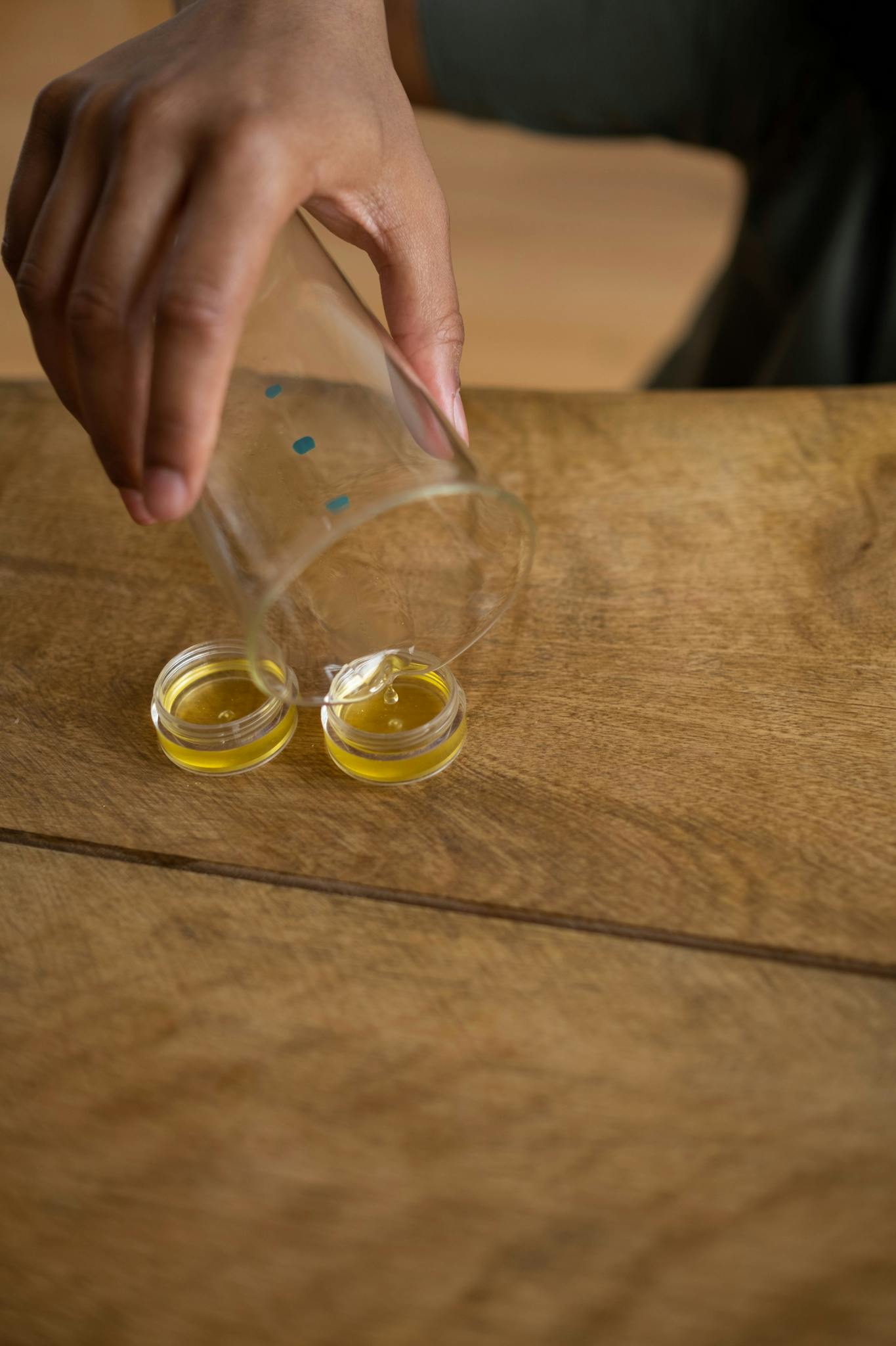 Close-up of a hand pouring yellow liquid into small containers on a wooden table.